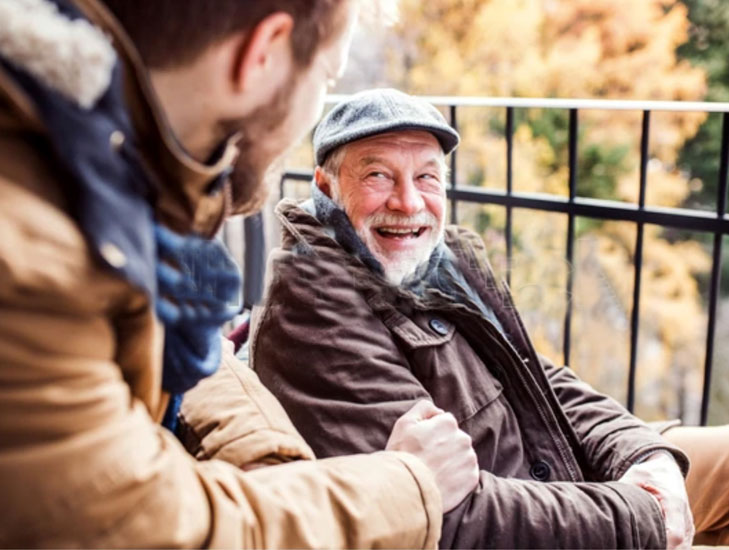 Mann sitzt neben freudig lächelndem älteren Herrn, herbstliche Bäume im Hintergrund.