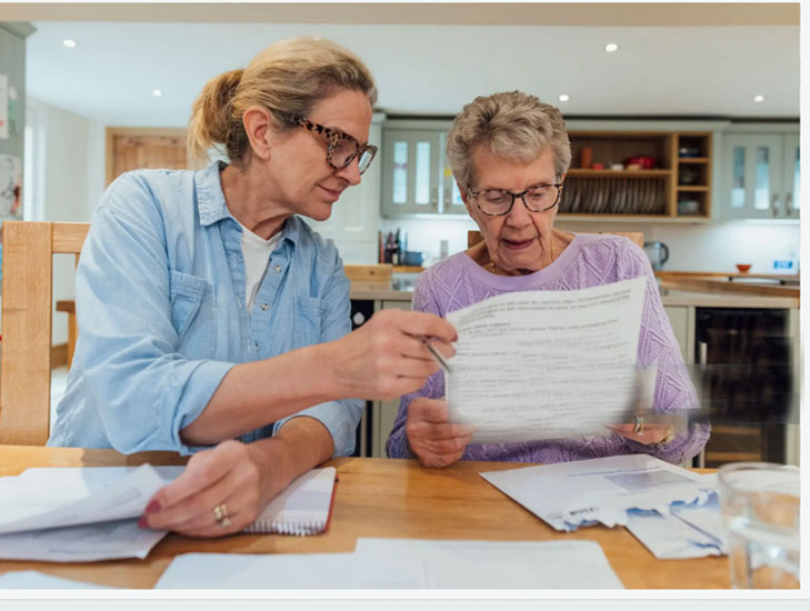 Jüngere Frau erklärt älterer Dame ein Formular, beide sitzen am Tisch mit zahlreichen Unterlagen und Papieren.