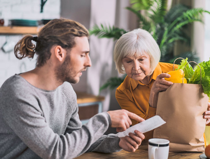 Ein junger Mann sitzt neben einer älteren Dame mit weißen Haaren, in häuslicher Situation. Er hält einen Zettel in der Hand und ist intensiv mit der Dame im Gespräch während sie eine Papier-Einkaufstüte vor sich stehend hat, aus der buntes Gemüse und Salat herausschauen.